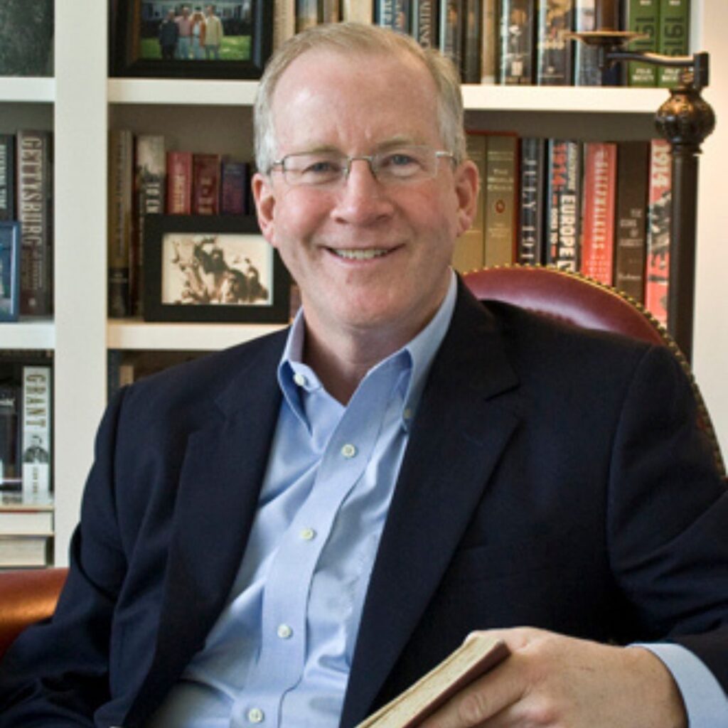 A man, Timothy Boyce, sitting in a chair holding a book in front a bookcase.