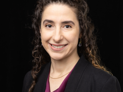 Woman in purple shirt and black blazer smiles against a black background