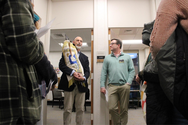 People gather in an office hallway as a bearded man holds a large wrapped package; staff and visitors watch, creating a friendly, busy mood.