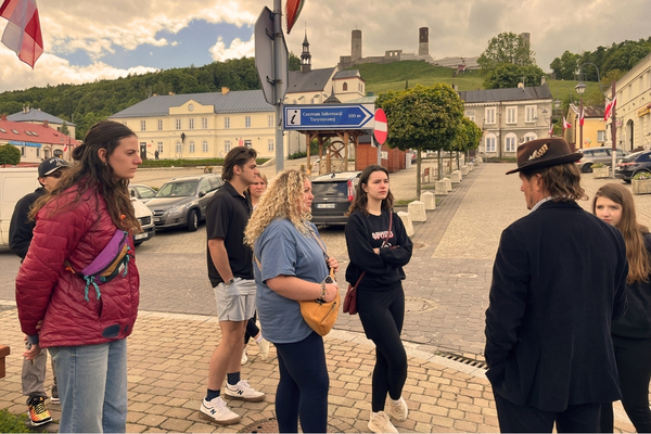 Tourists gather in a historic European square with a guide, colorful flags, cobblestones, ornate buildings, and a cloudy sky.