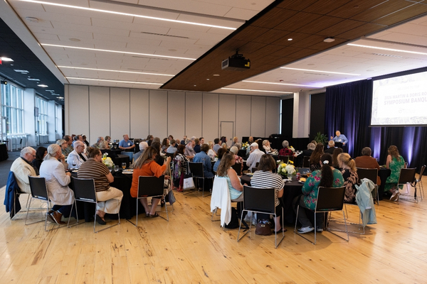 Conference attendees gather around round tables in a bright event hall, listening to a presentation on a lit screen; engaged, collaborative atmosphere.