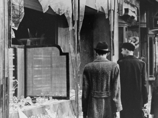 Black-and-white wartime street scene: two men walk past a bomb-damaged shop with shattered windows.